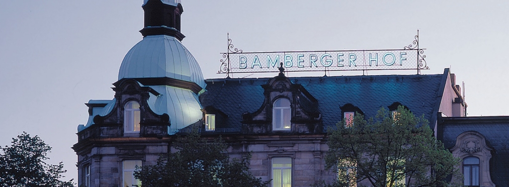 Hotel Bamberger Hof: Historisches Gebäude mit Kuppeldach, großes Schild "Bamberger Hof", Bäume in der Abenddämmerung.