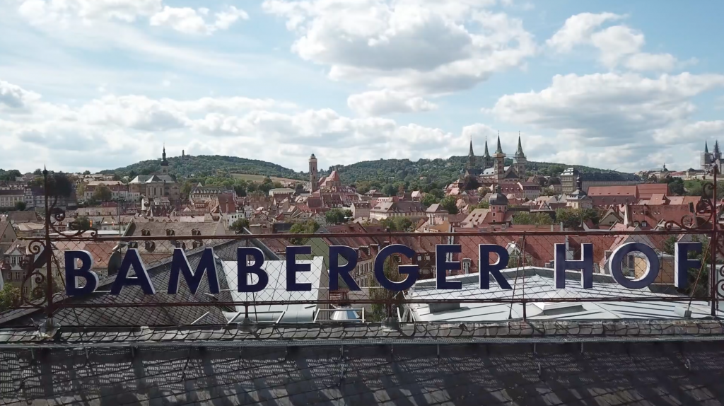Dachschild "Hotel Bamberger Hof" mit Blick auf die Bamberger Altstadt, rote Dächer, Kirchtürme, Hügel, bewölkter Himmel.