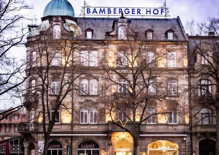Hotel Bamberger Hof: Historisches Gebäude, beleuchtete Fenster, blauer Kuppelturm, blattlose Bäume, Menschen in Rot auf Bänken in der Abenddämmerung.