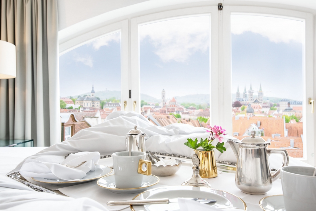 Ein Frühstückstablett mit Kaffee und Blumen auf einem Bett im Hotel Bamberger Hof, vor großen Fenstern mit Blick auf das historische Stadtbild.