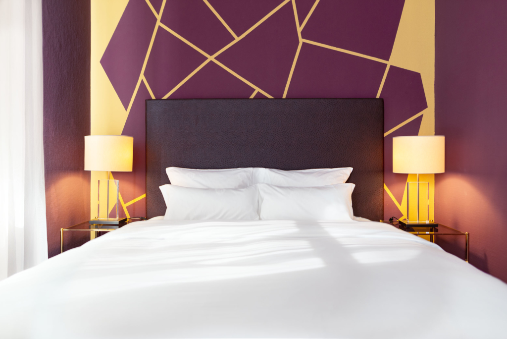 Ein ordentlich gemachtes Bett mit weißer Bettwäsche, zwei Nachttische mit Lampen, eine lila und gelbe geometrische Akzentwand im Hotel Bamberger Hof.