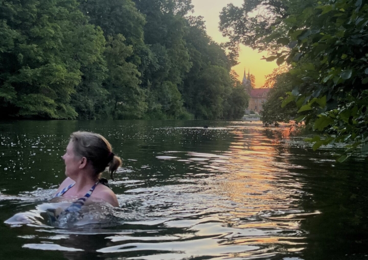 Eine Person mit geflochtenen Haaren schwimmt in einem ruhigen Fluss in der Nähe des Hotels Bamberger Hof, umgeben von dichtem, grünem Baumbewuchs bei Sonnenuntergang.