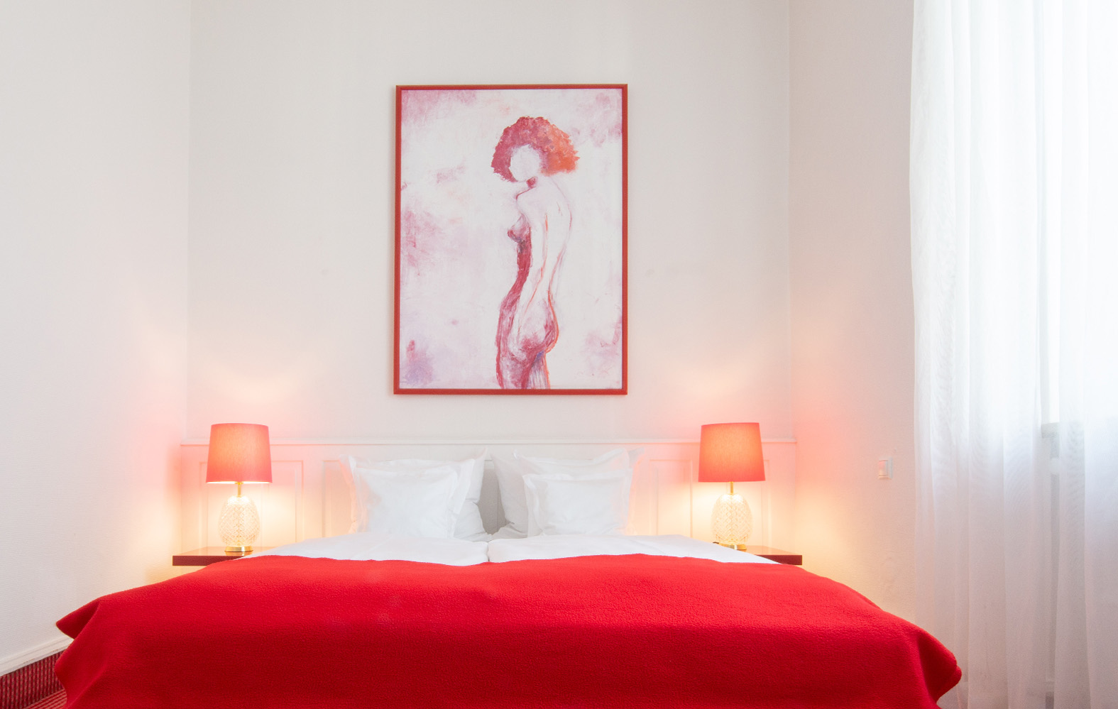 Im Hotel Bamberger Hof: Ein Bett mit roter Decke, weiße Kissen, zwei beleuchtete Lampen, ein Aktgemälde über dem Kopfteil.